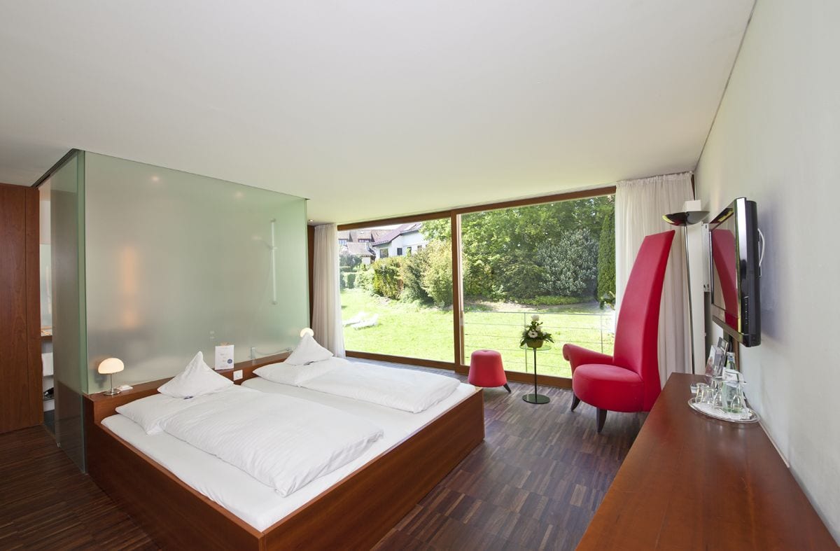 Habitación de hotel moderna con una cama grande, un televisor montado en la pared y vistas al jardín.