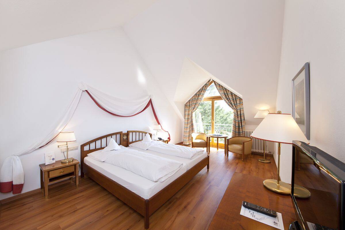 Un dormitorio con una cama grande, una mesita de noche, una lámpara, una silla, un televisor y un ventanal con cortinas.