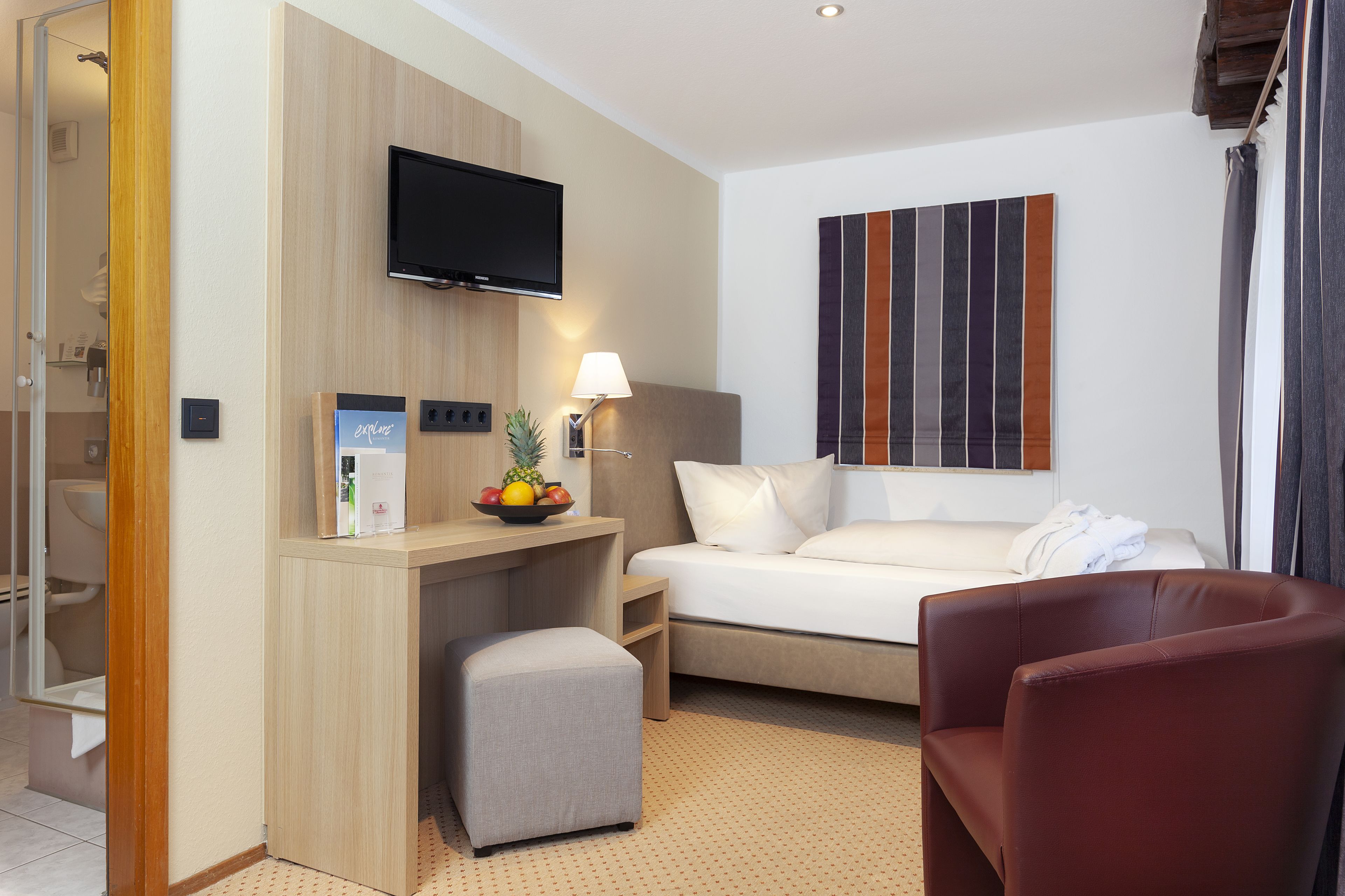 Een hotelkamer met een bed, een bureau met een computer, een televisie, een lamp, een schaal fruit, en een rode stoel.