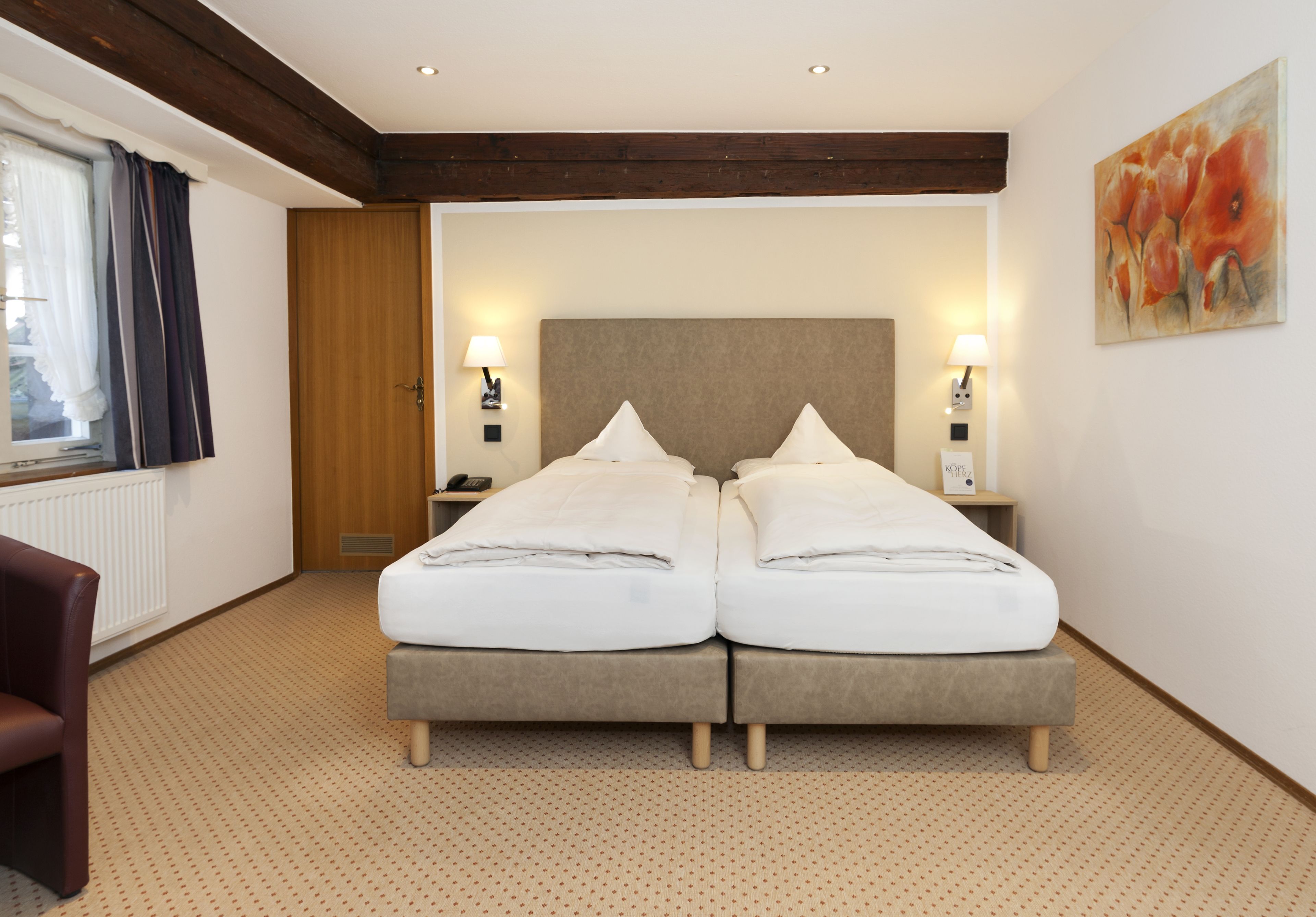 Habitación de hotel con una cama doble, dos mesitas de noche con lámparas, una puerta de madera, una ventana con cortinas y un cuadro en la pared.