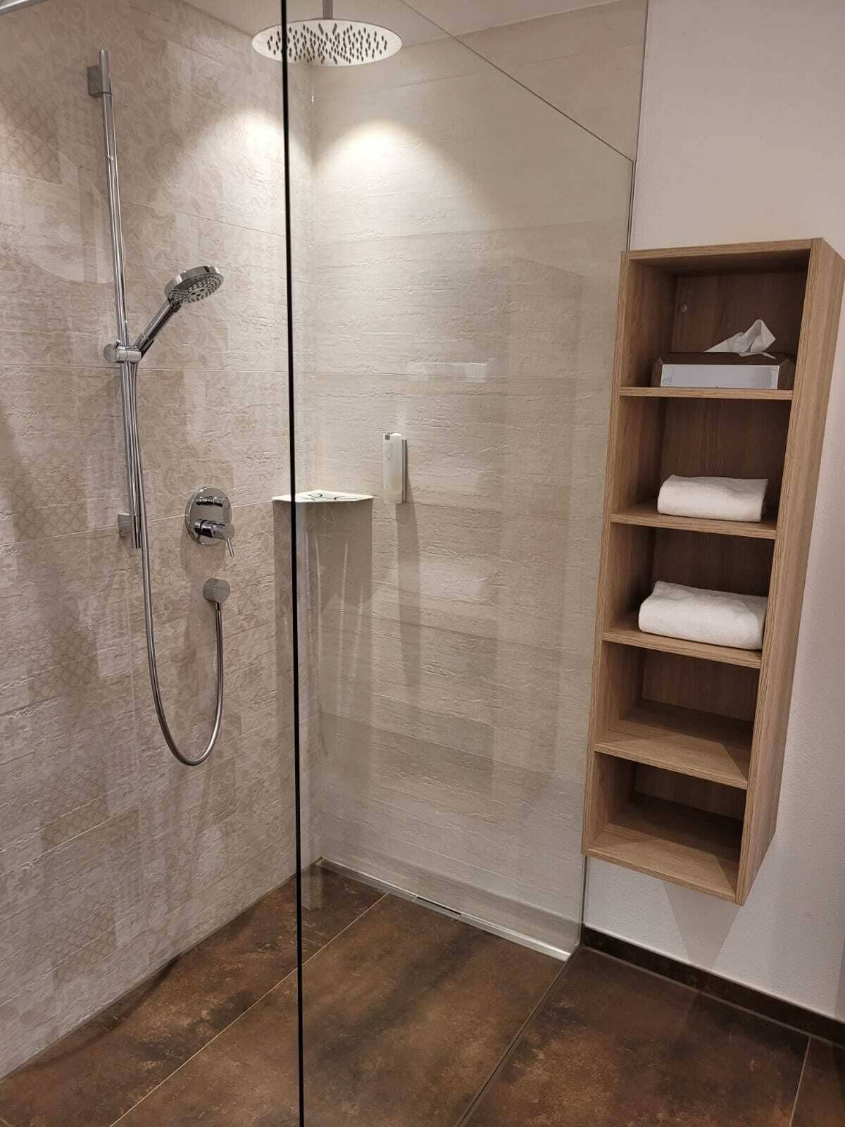 A modern bathroom with a glass shower enclosure, a wooden shelf with towels, and a marble-tiled floor.