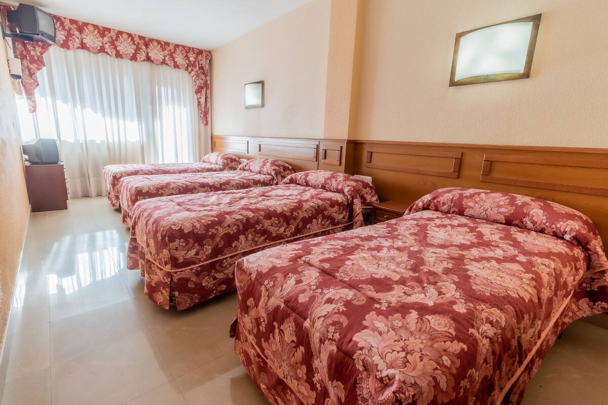 Habitación de hotel con tres camas, televisión, espejo y ventana con cortinas.