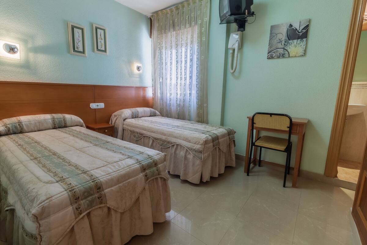 Habitación de hotel con dos camas, una mesita de noche de madera, una silla, un teléfono y un cuadro en la pared.