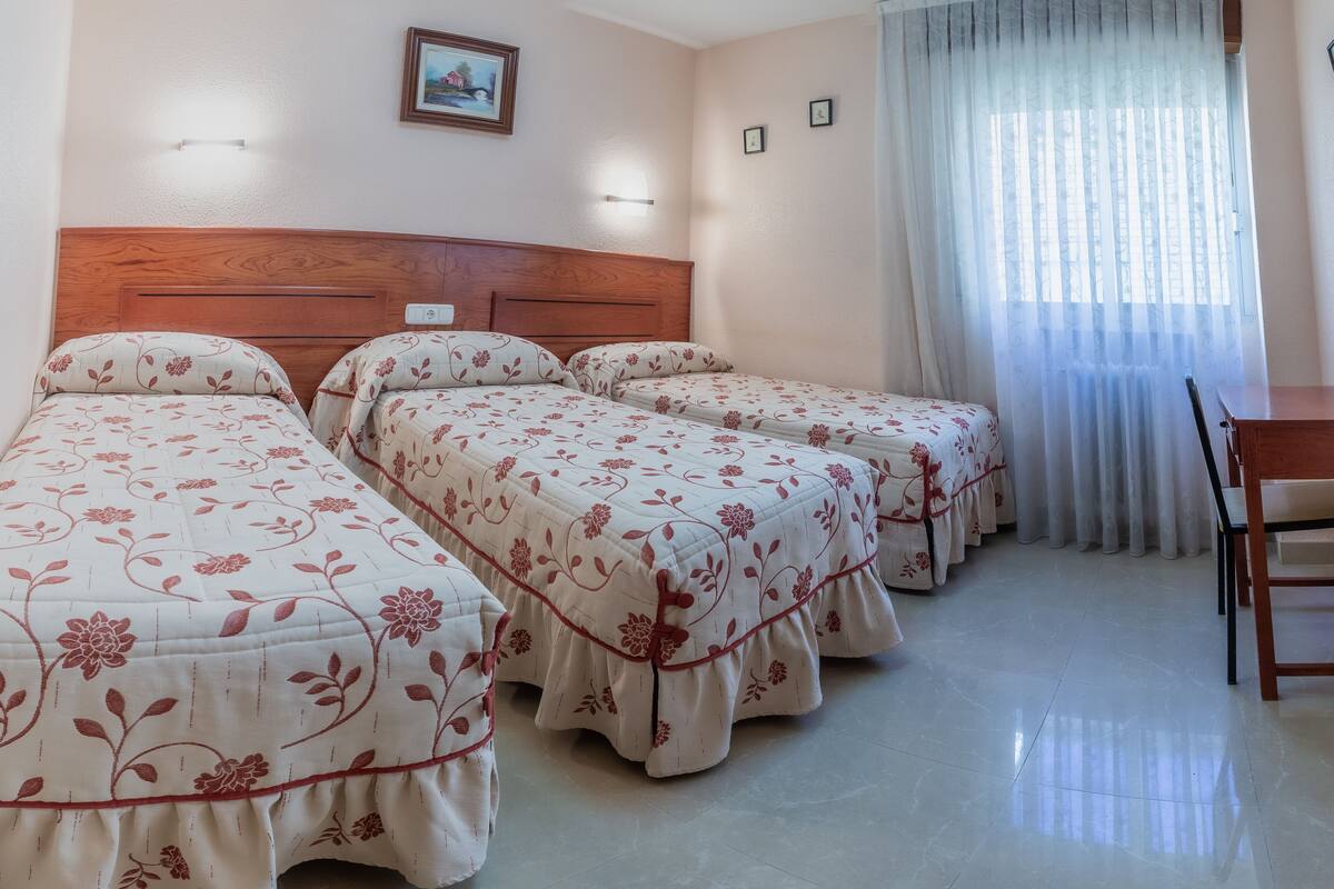 Habitación de hotel con tres camas individuales, cabecera de madera, una mesita y una ventana con cortinas traslúcidas.