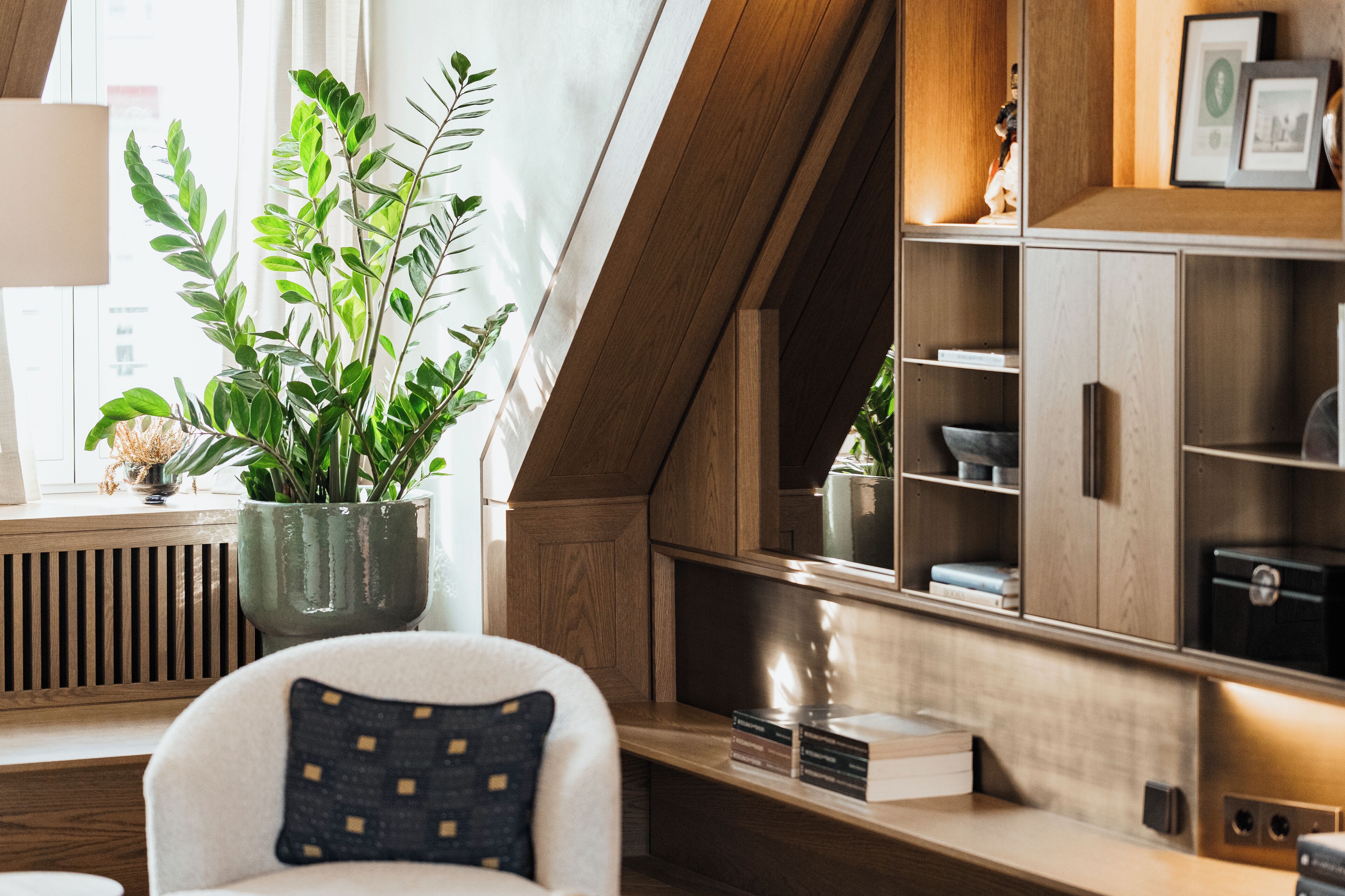 A modern living room with a wooden sloped ceiling, a large potted plant, a white armchair with a patterned pillow, and built-in wooden shelves with various items.