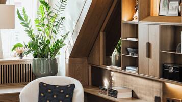 A modern living room with a wooden sloped ceiling, a large potted plant, a white armchair with a patterned pillow, and built-in wooden shelves with various items.