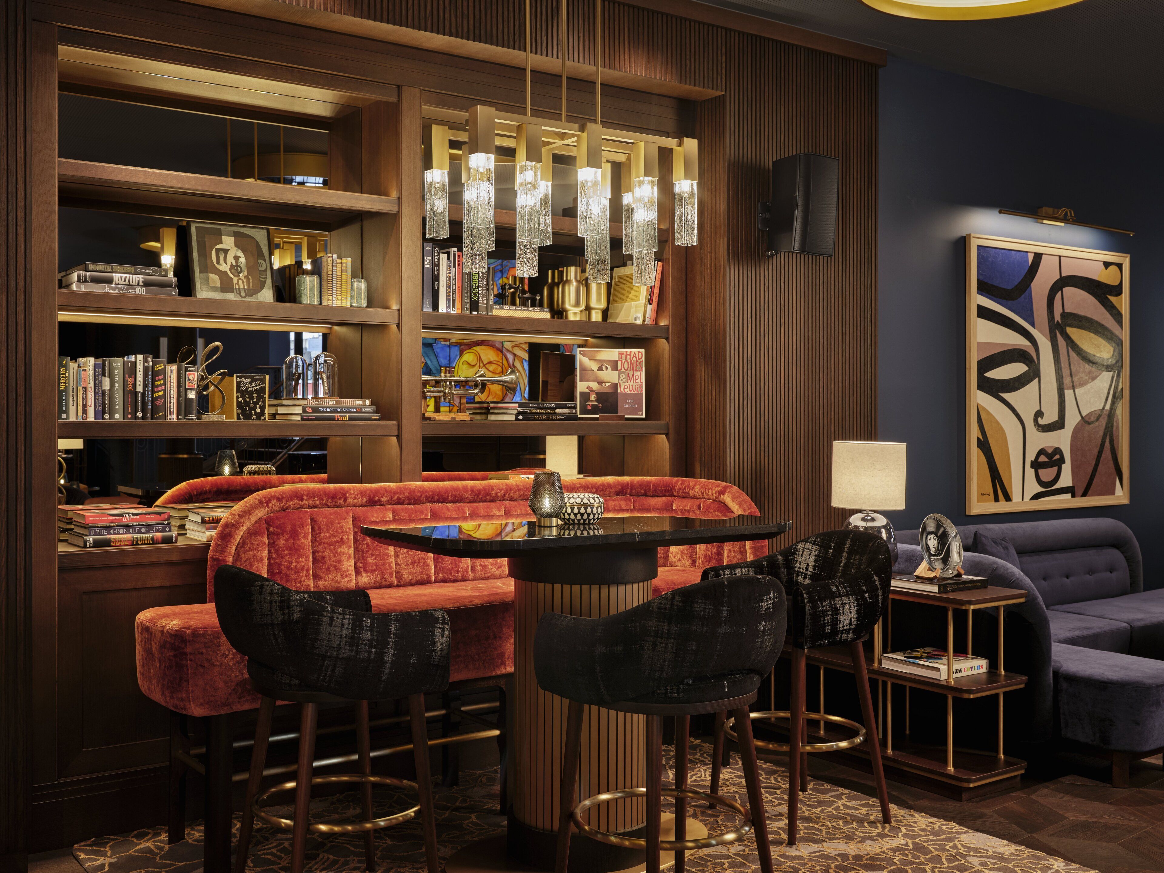 A modern bar area with a red cushioned bench, black chairs, and a small round table. There are bookshelves filled with books and decorative items, and a large abstract painting on the wall.