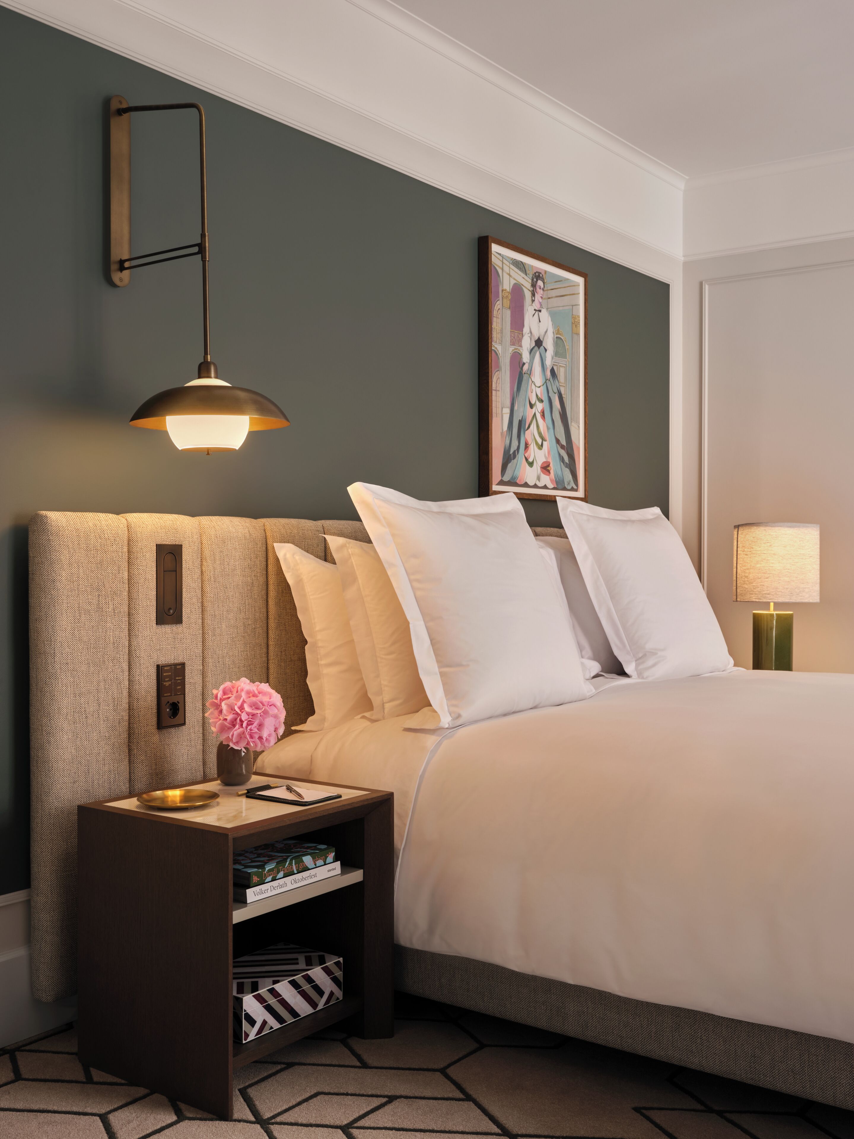 Una cama bien hecha con ropa de cama blanca, una mesita de noche con un florero y una lámpara de pared, además de un cuadro enmarcado sobre la cama.