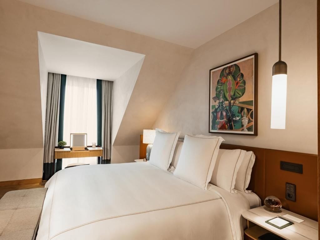 Habitación de hotel con una cama grande, un escritorio con silla, una mesita de noche, una lámpara y un cuadro en la pared.