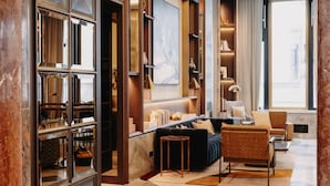 A modern living room with a sofa, armchairs, and a coffee table. There are bookshelves and a large window with a view of the city. - Rosewood Munich (Munich)