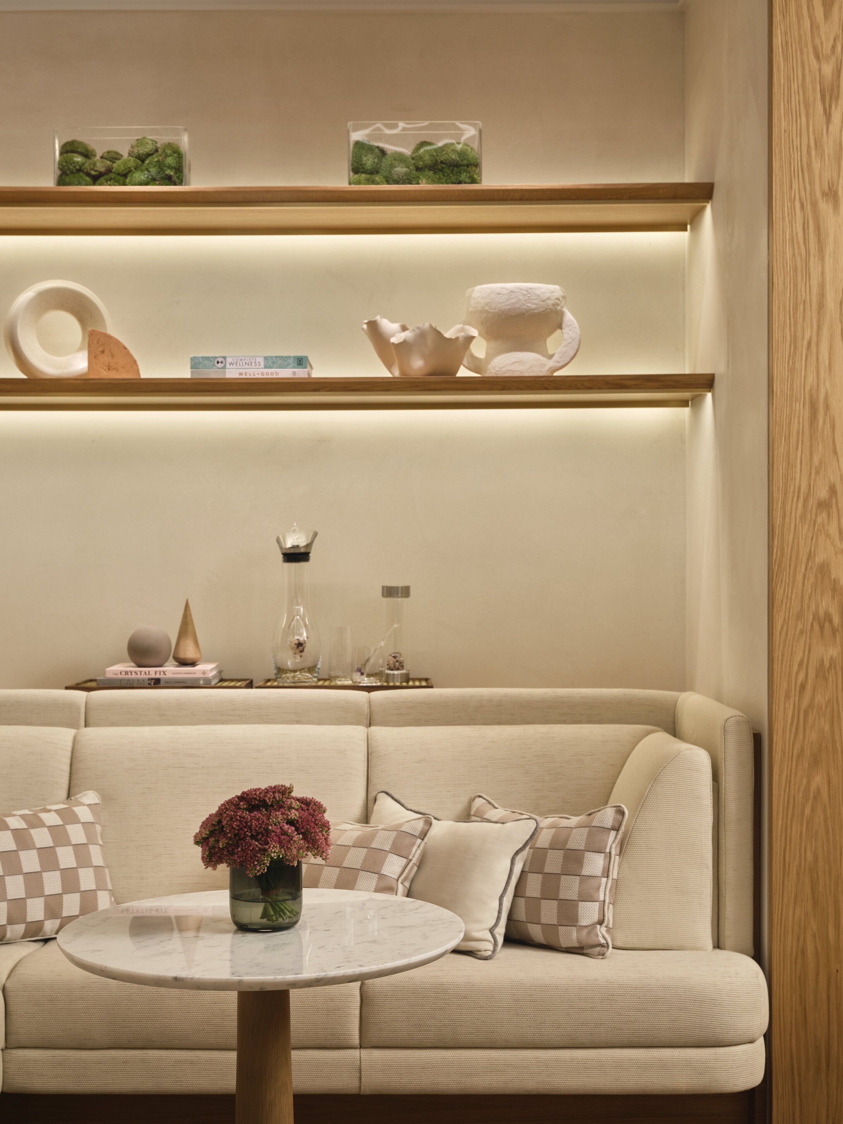 Un salon moderne avec un canapé beige, une table basse ronde en marbre et des objets décoratifs disposés sur des étagères au-dessus.
