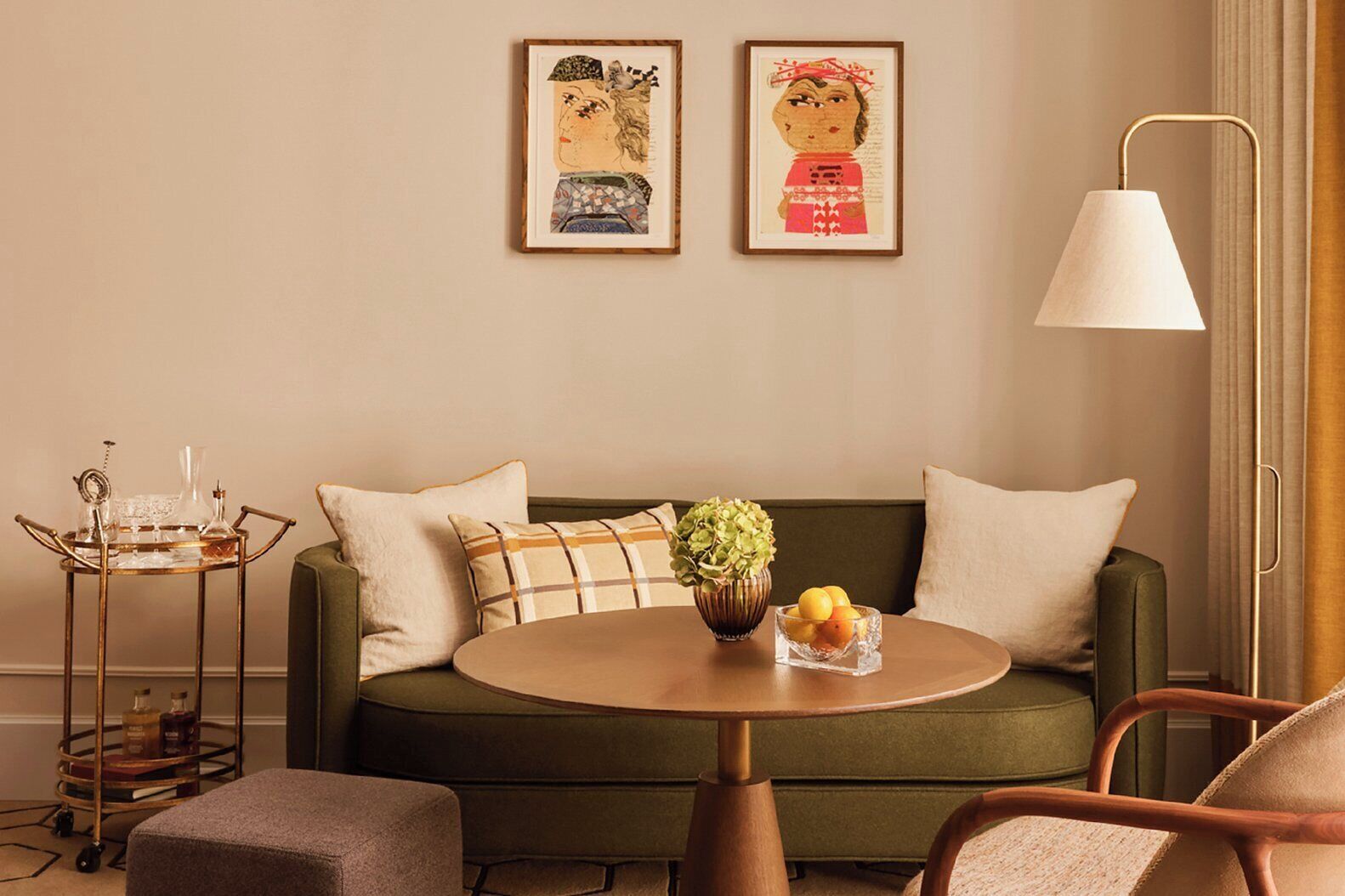 Una sala de estar acogedora con un sofá verde, una mesa redonda con fruteros, una mesita de noche con lámpara y dos cuadros en la pared.