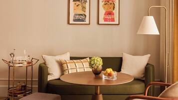 Una sala de estar acogedora con un sofá verde, una mesa redonda con fruteros, una mesita de noche con lámpara y dos cuadros en la pared.