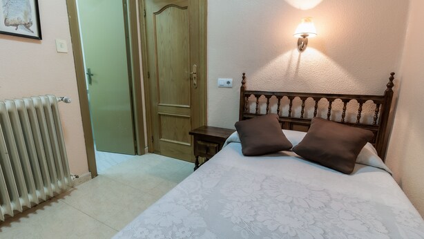 Un dormitorio con una cama, dos almohadas, una mesita de noche, una lámpara de pared, un radiador y una puerta.