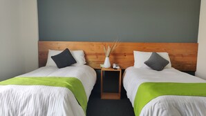 Ein Hotelzimmer mit zwei Einzelbetten, jeweils mit Holz Kopfteil, weißer Bettwäsche und grünen Tagesdecken. Ein Nachttisch zwischen den Betten beherbergt eine Vase mit Trockenblumen und ein Telefon.
