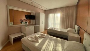 Habitación de hotel con cama, mesita de noche, un escritorio pequeño con silla, televisor montado en la pared y ventana con cortinas.