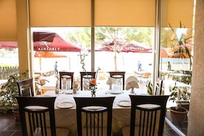 An indoor dining area with tables set for four, chairs, and a view of the outside through large windows.