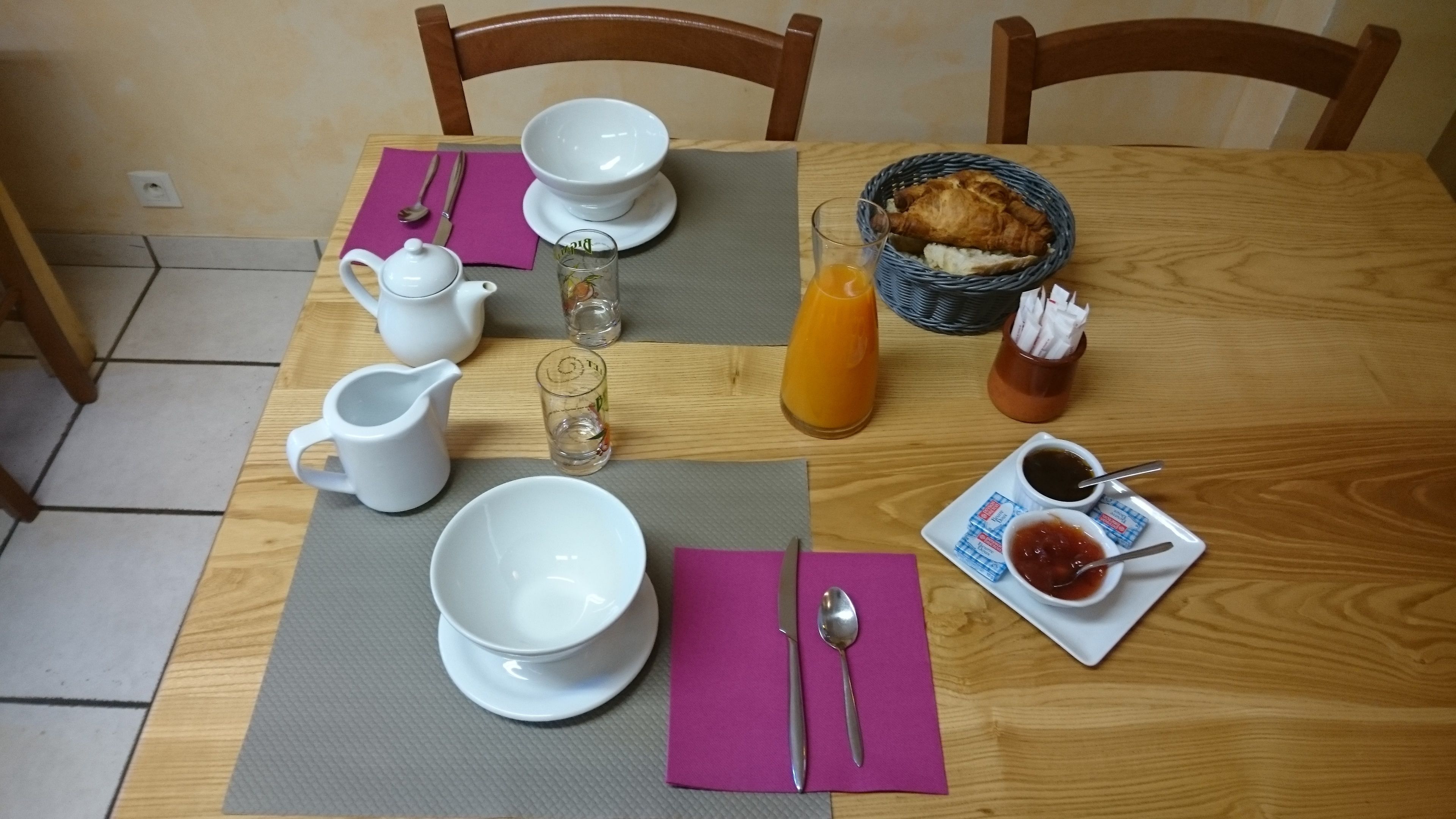 Une table de petit-déjeuner dressée avec de la vaisselle blanche, une théière, un verre de jus d’orange, un panier de croissants et des condiments.