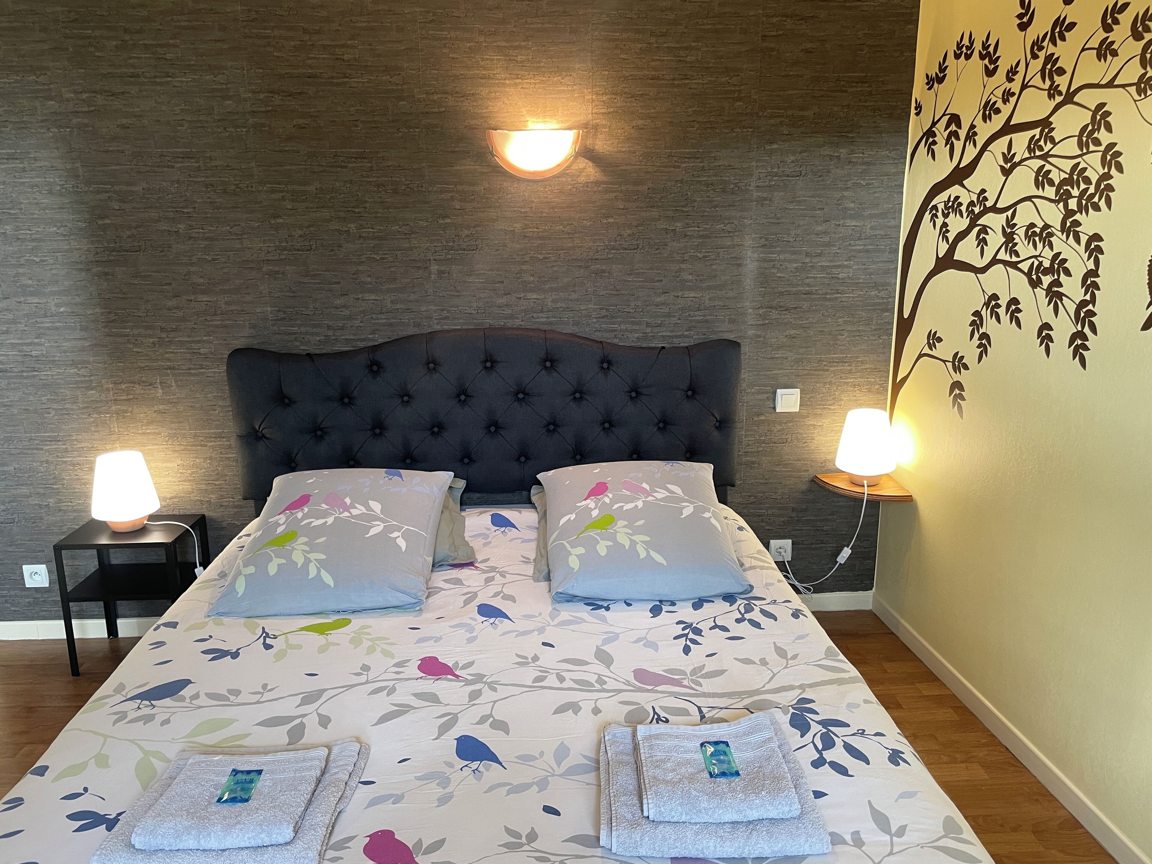 A neatly made bed with floral bedding and two pillows, flanked by bedside lamps on black nightstands, against a textured wall with a tree mural.