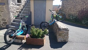 Deux vélos sont garés dans une allée, devant un mur de pierres et un bac à fleurs en bois où sont plantées des plantes.
