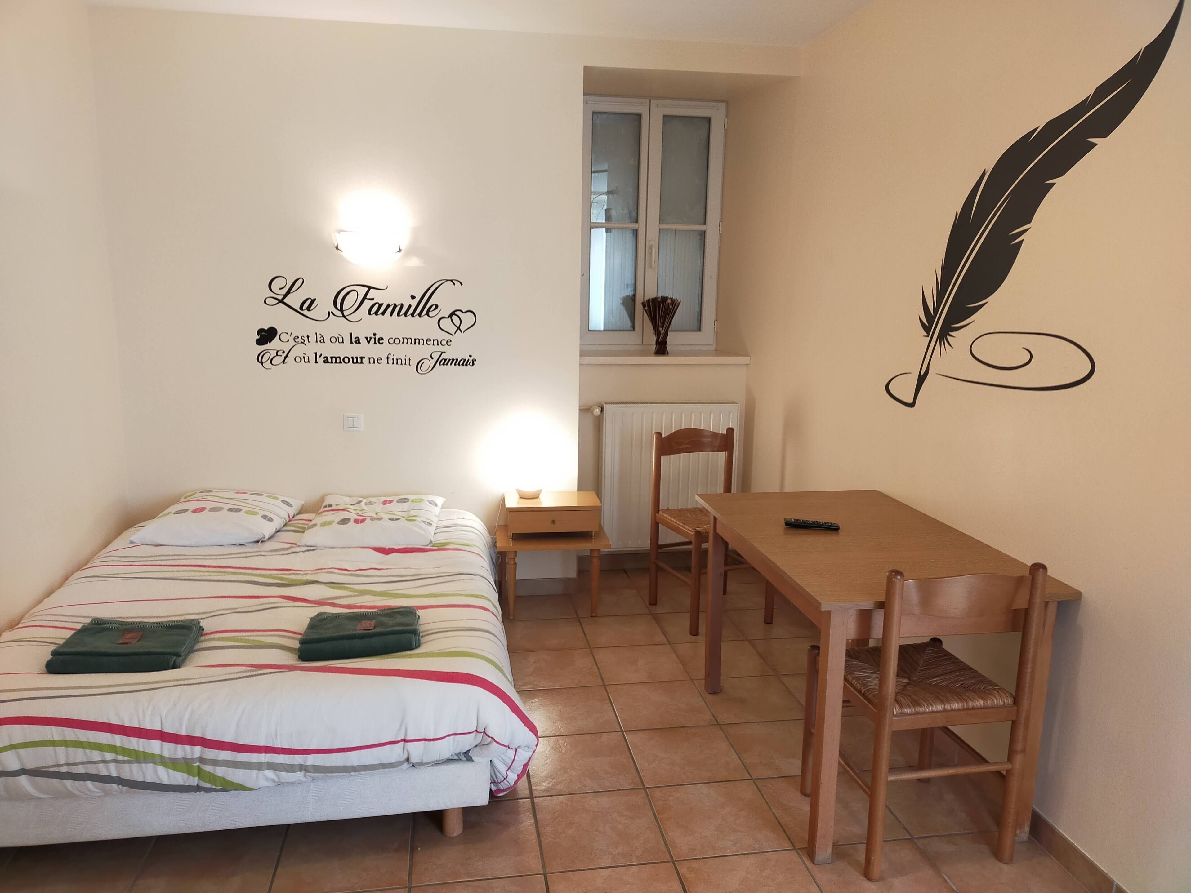 A bedroom with a bed, a wooden table, and a chair. There is a wall decoration with a feather and a sign that says "La Famille".