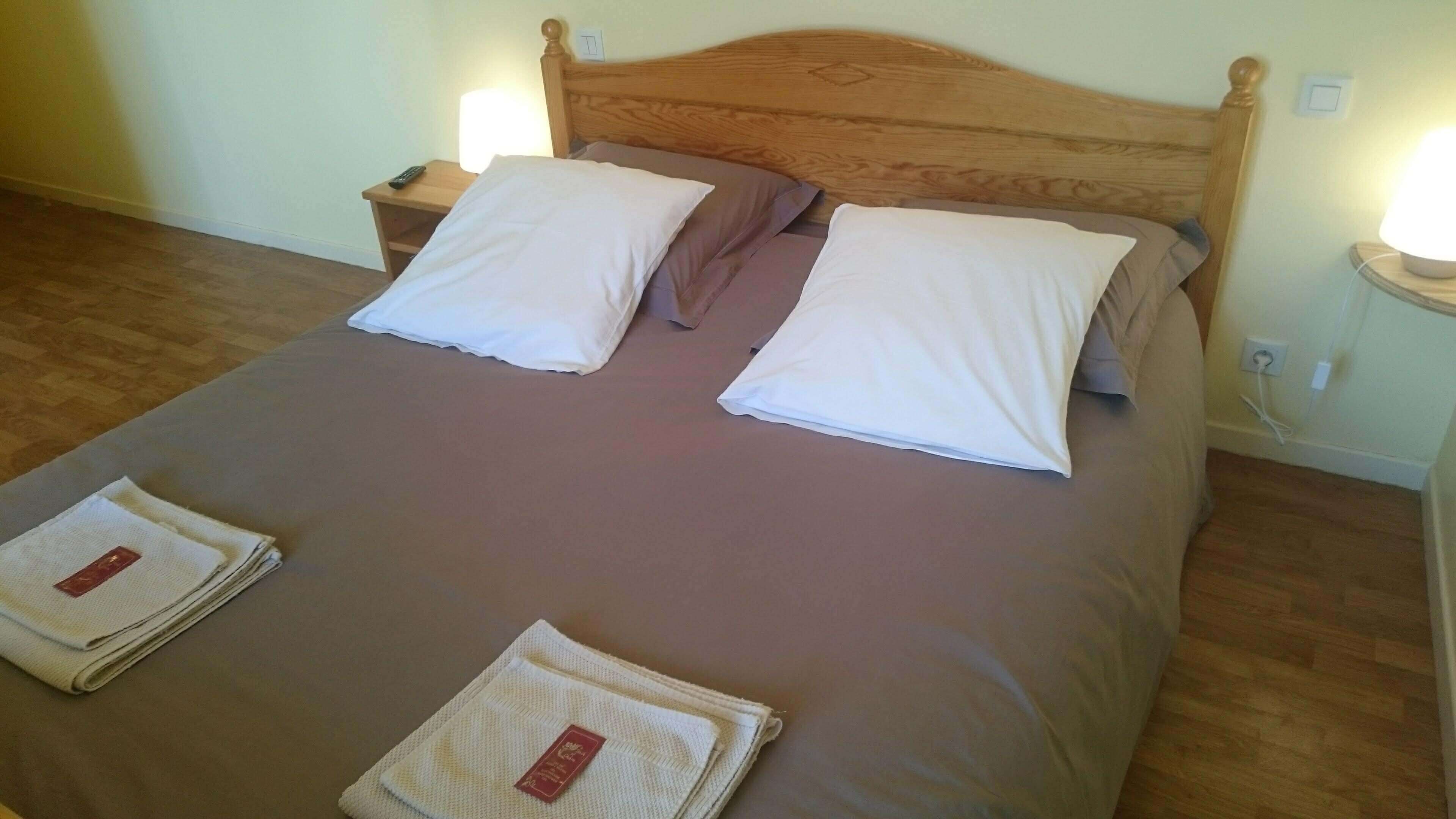 Una cama bien hecha con cabecera de madera, dos almohadas blancas y una colcha marrón. Hay dos mesitas de noche con lámparas, y una toalla doblada sobre la cama.