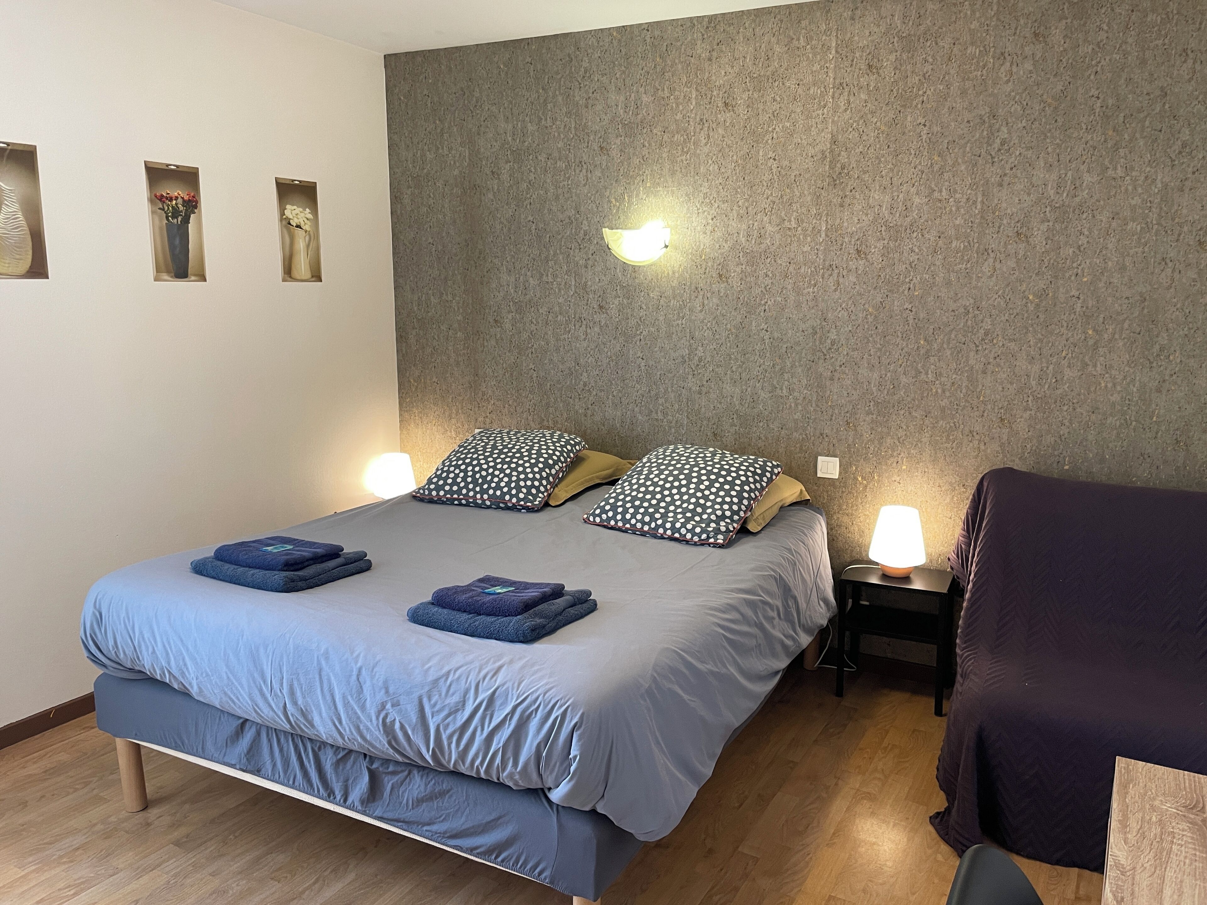 A neatly arranged bedroom with a bed, two bedside tables with lamps, a wall-mounted light, and a wooden floor.
