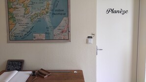 Une carte accrochée au mur, une porte portant l’inscription « Planée », et un bureau en bois avec un tableau noir et du matériel d’écriture.