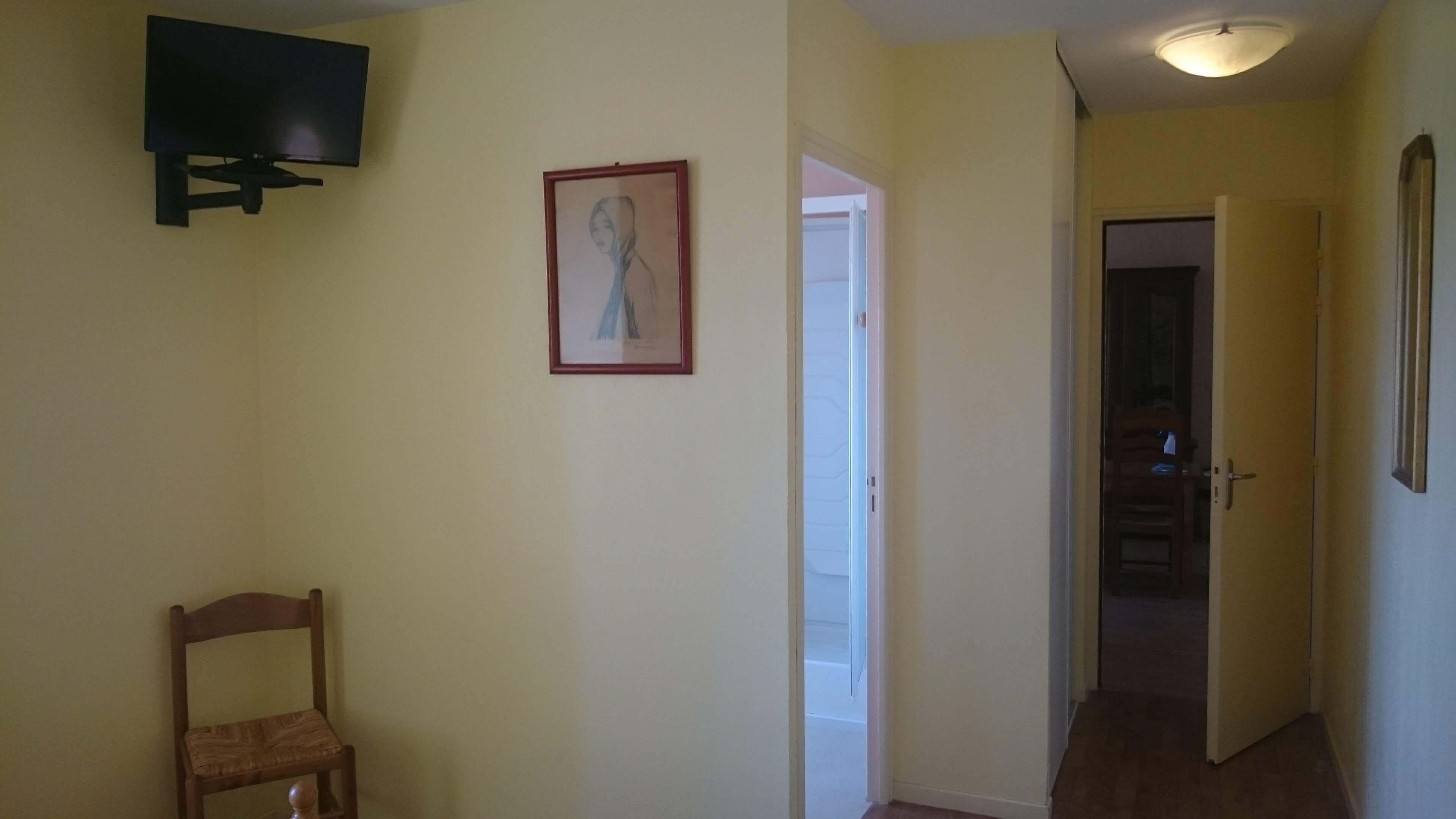 Een gang met een houten stoel, een ingelijst kunstwerk aan de muur en een aan de muur bevestigde televisie.