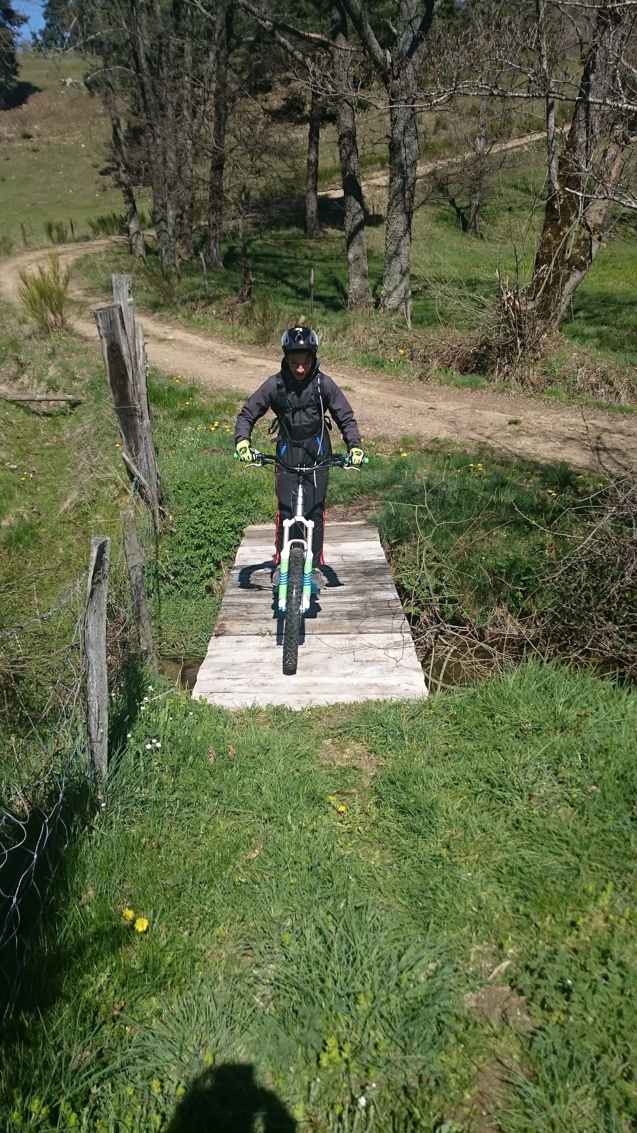 Een persoon fietst over een houten brug in een natuurlijke omgeving met bomen en gras.