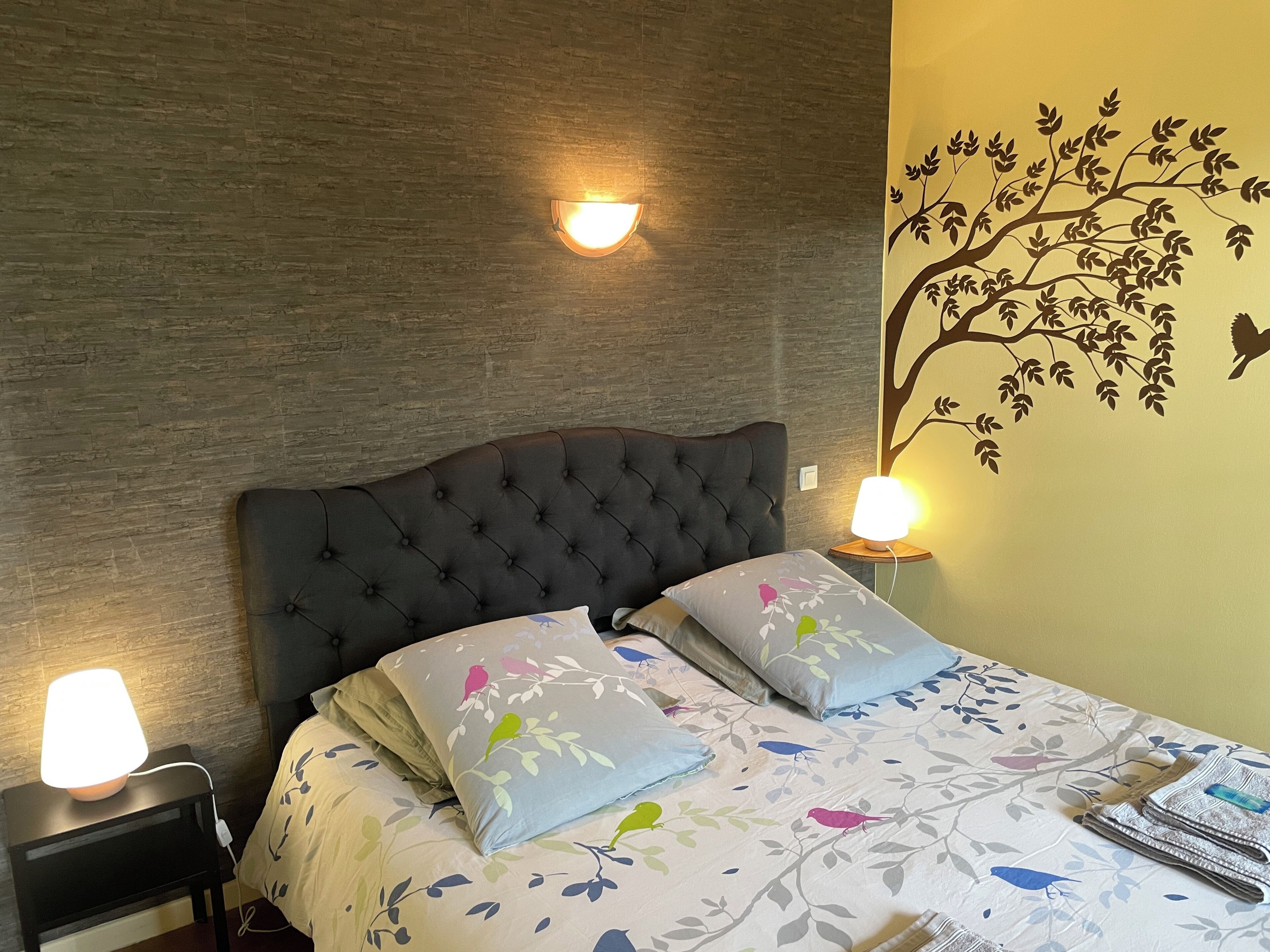 A bedroom with a tufted headboard, two bedside lamps, a bed with floral-patterned bedding, and a wall mural of a tree with birds.