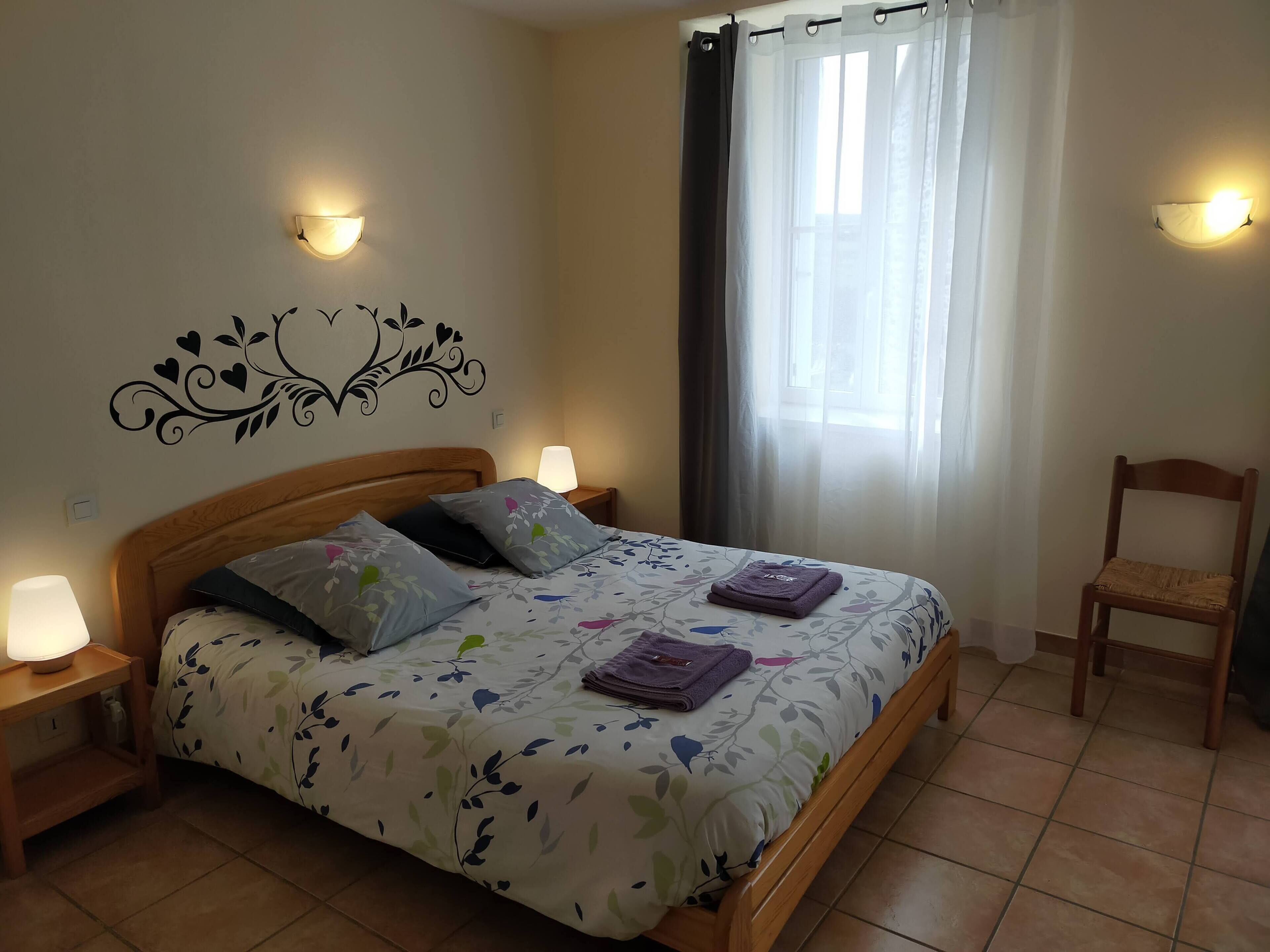 Un lit bien fait, avec une literie à motifs floraux, deux lampes de chevet, un autocollant décoratif sur le mur, une chaise et une fenêtre avec des rideaux légers.