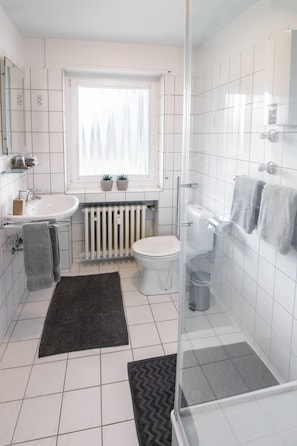 A modern bathroom with a white sink, toilet, and shower. There are two potted plants on the windowsill, a towel rack with towels, and a black rug on the floor.