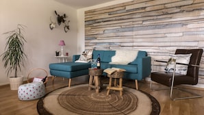 Ein Wohnzimmer mit einem blauen Sofa, einem runden Teppich, einem Holztisch, einem Topfpflanzen und einer Wand mit Holzbodenoptik.