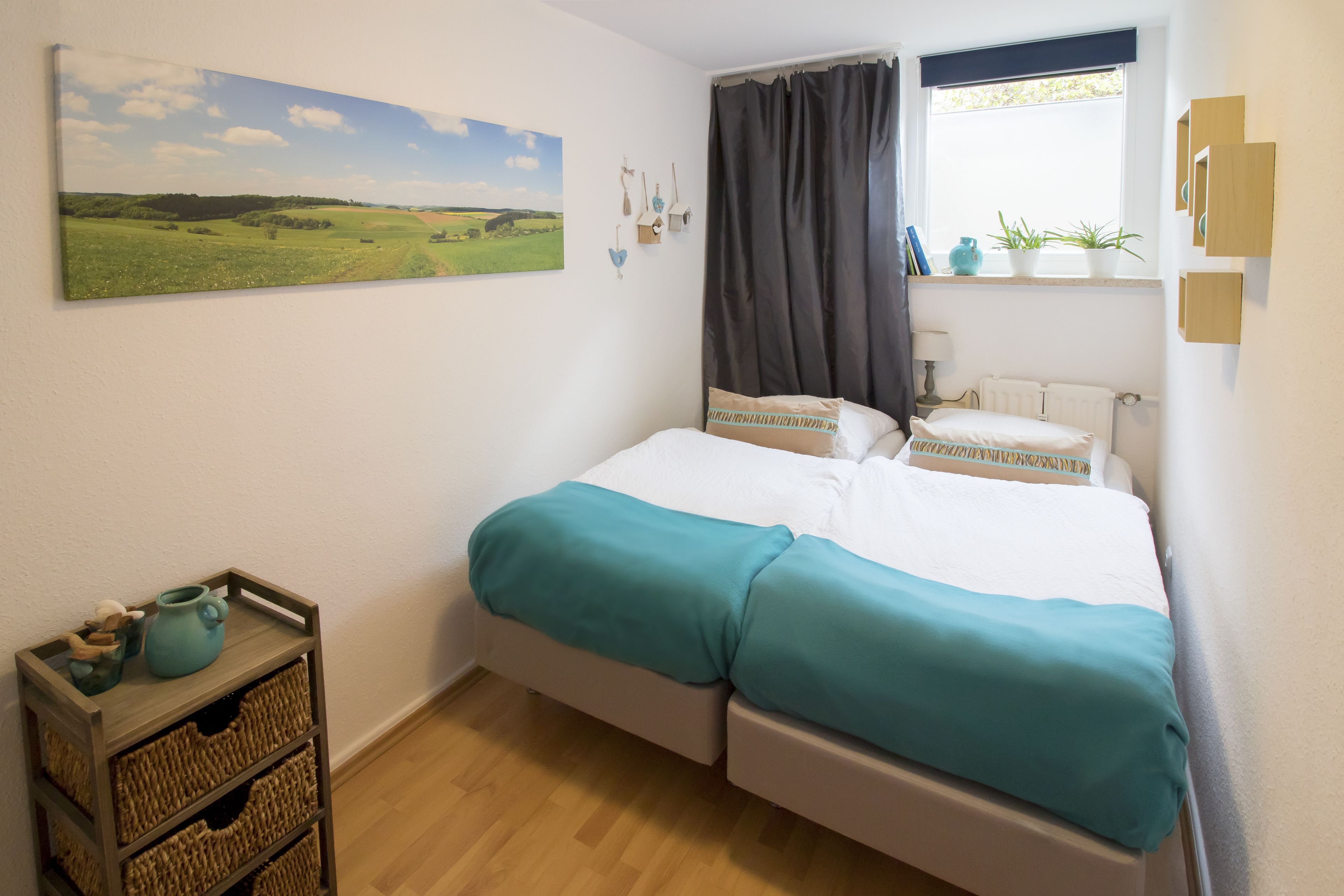 Un dormitorio con una cama, una ventana con cortinas, un cuadro en la pared y una mesita con un jarrón y una cesta.
