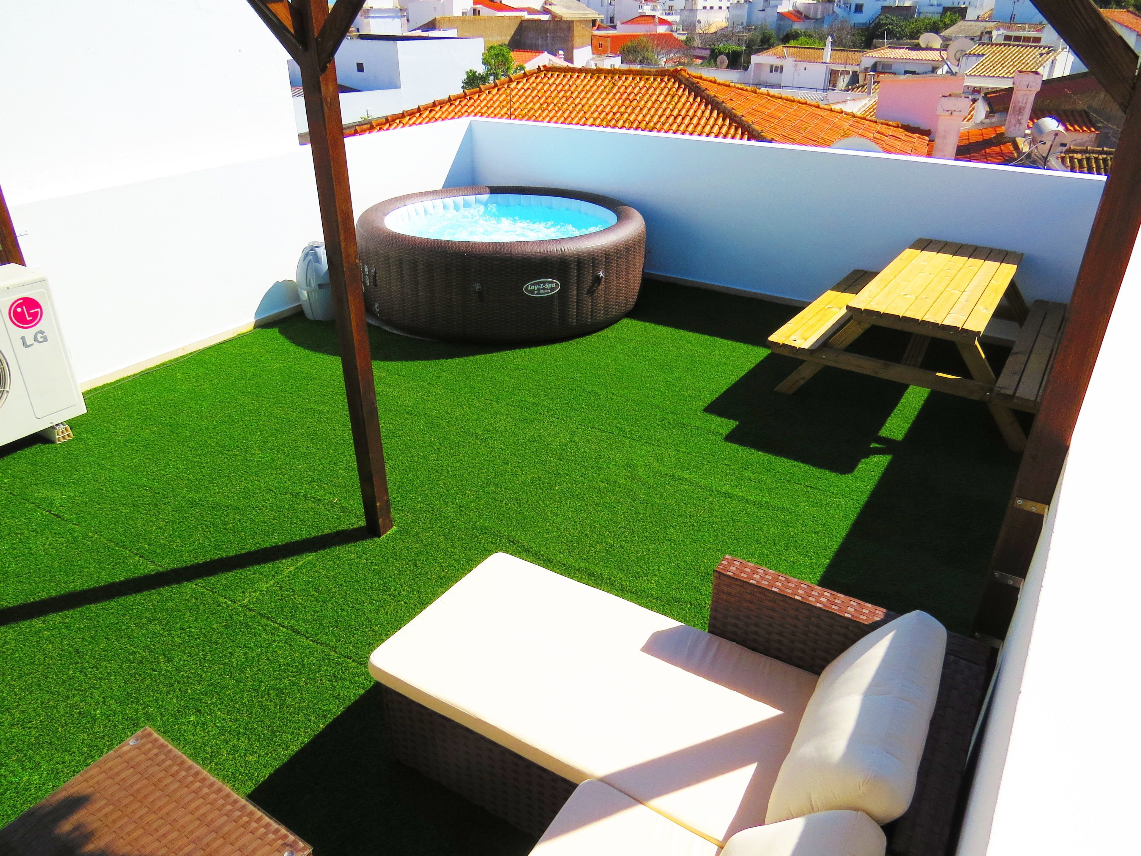 A rooftop patio with artificial grass, a hot tub, a wooden table, and a lounge chair.