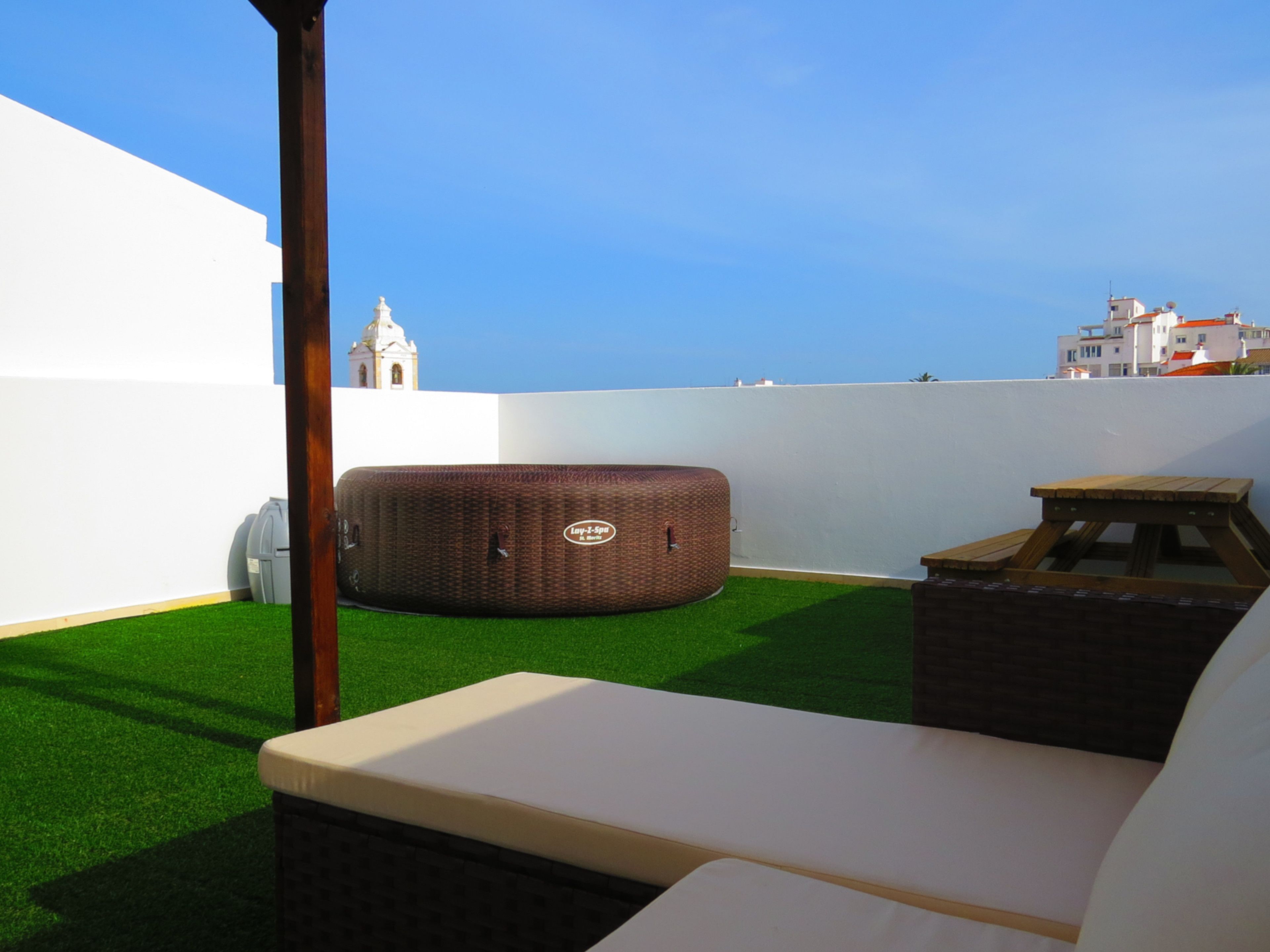 A rooftop terrace with artificial grass, a wicker ottoman, a wooden table, and a white wall.