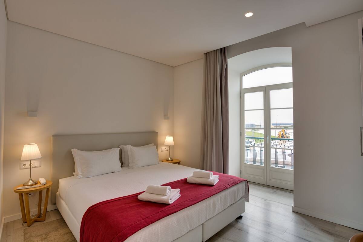 A hotel room with a large bed, two bedside lamps, a red throw blanket, and a balcony with a view.