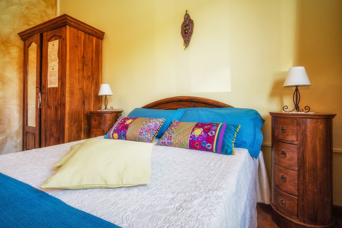 Un lit bien fait, avec une couverture bleue et des coussins colorés, une armoire en bois et deux tables de chevet avec des lampes.
