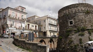 Una torre de piedra histórica con parte superior circular, junto a una hilera de edificios tradicionales con balcones y señalización de negocios locales.