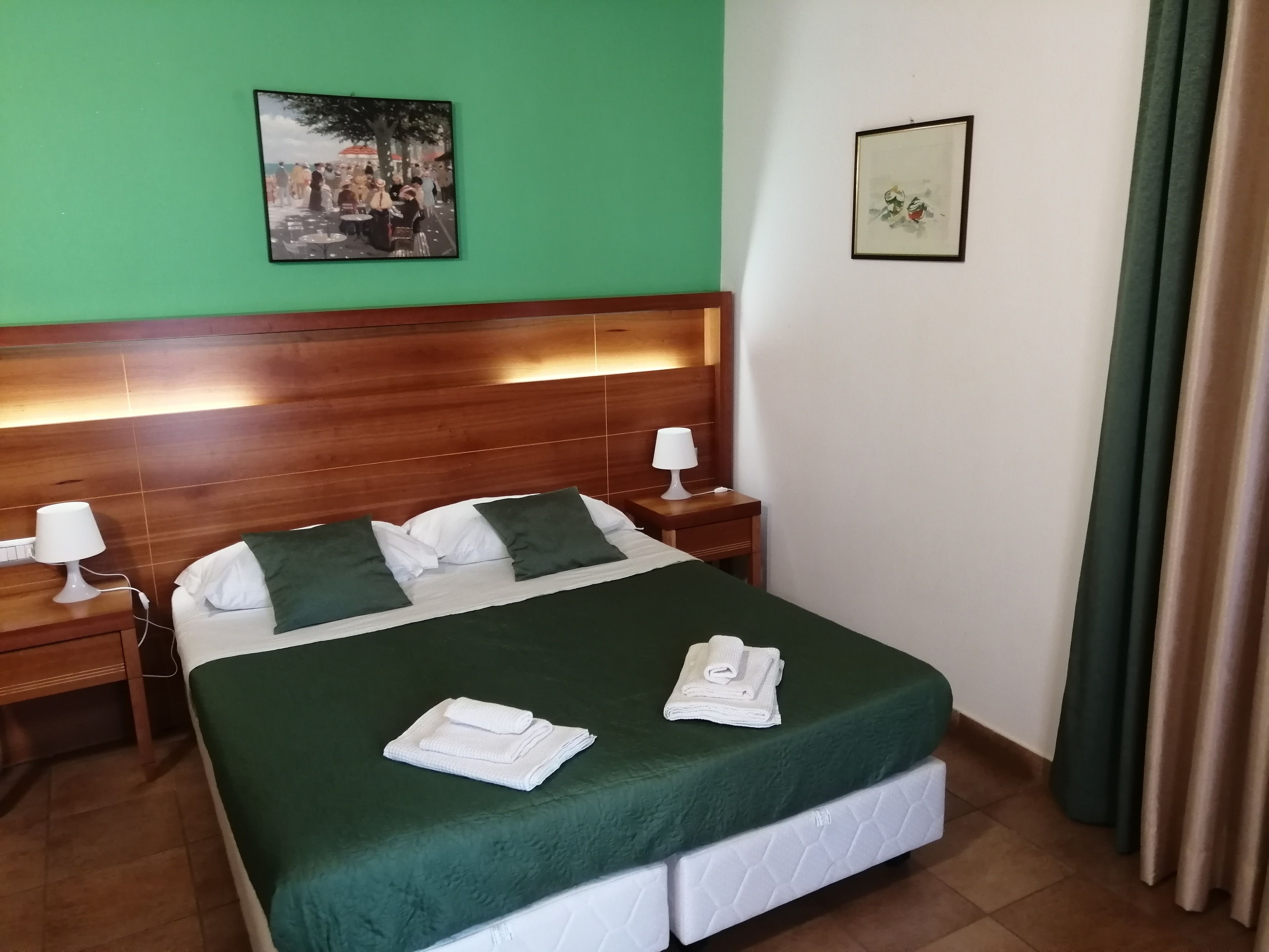 A hotel room with a wooden headboard, a green bedspread, two pillows, a white blanket, and a folded towel on the bed. There is a framed picture on the wall and a nightstand with a lamp.