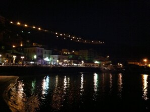Vista nocturna de un pueblo costero con edificios a lo largo del muelle, iluminados por farolas y reflejados en el agua.
