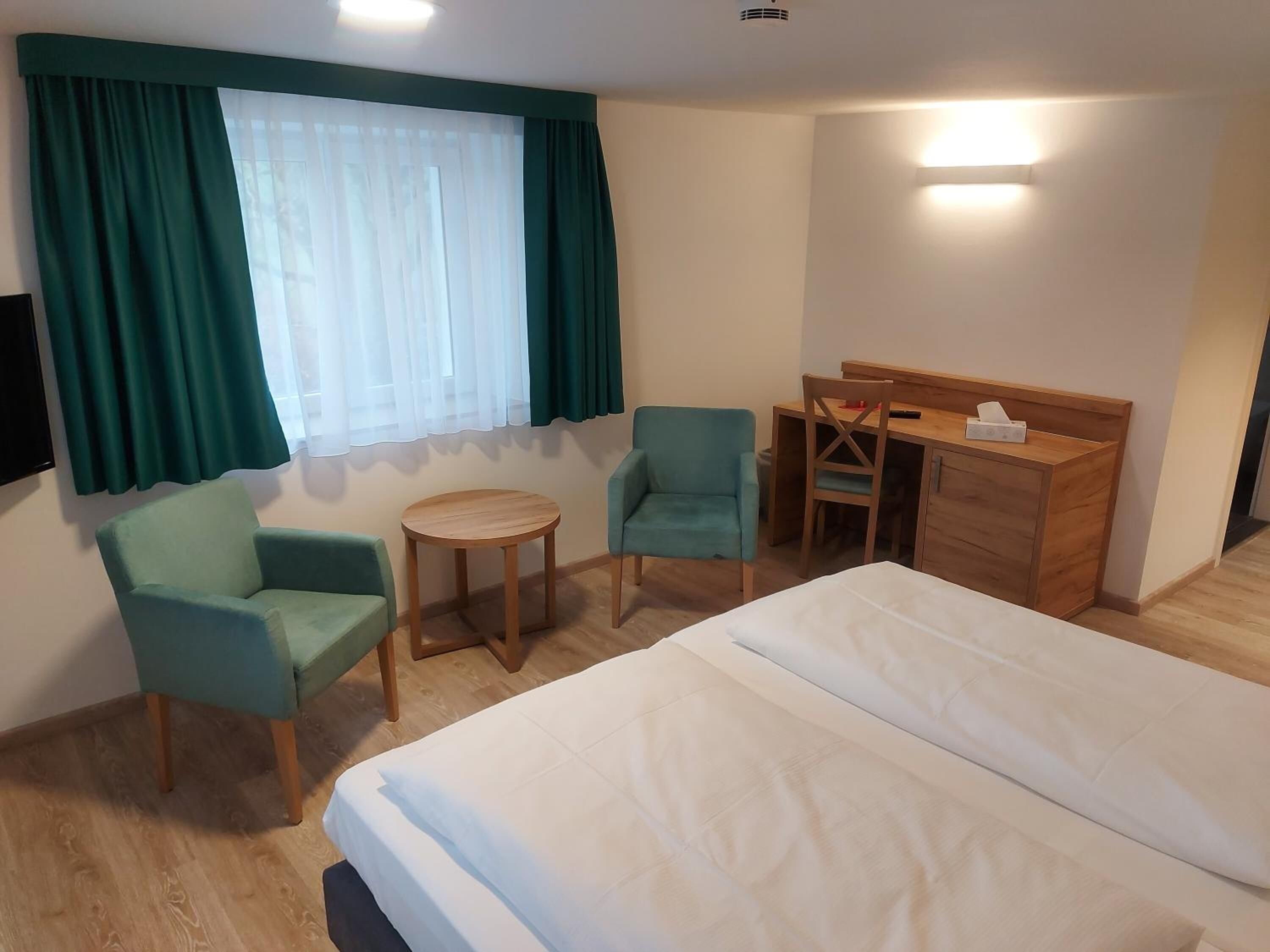 A hotel room with a bed, two armchairs, a small round table, a desk with a chair, and a window with curtains.