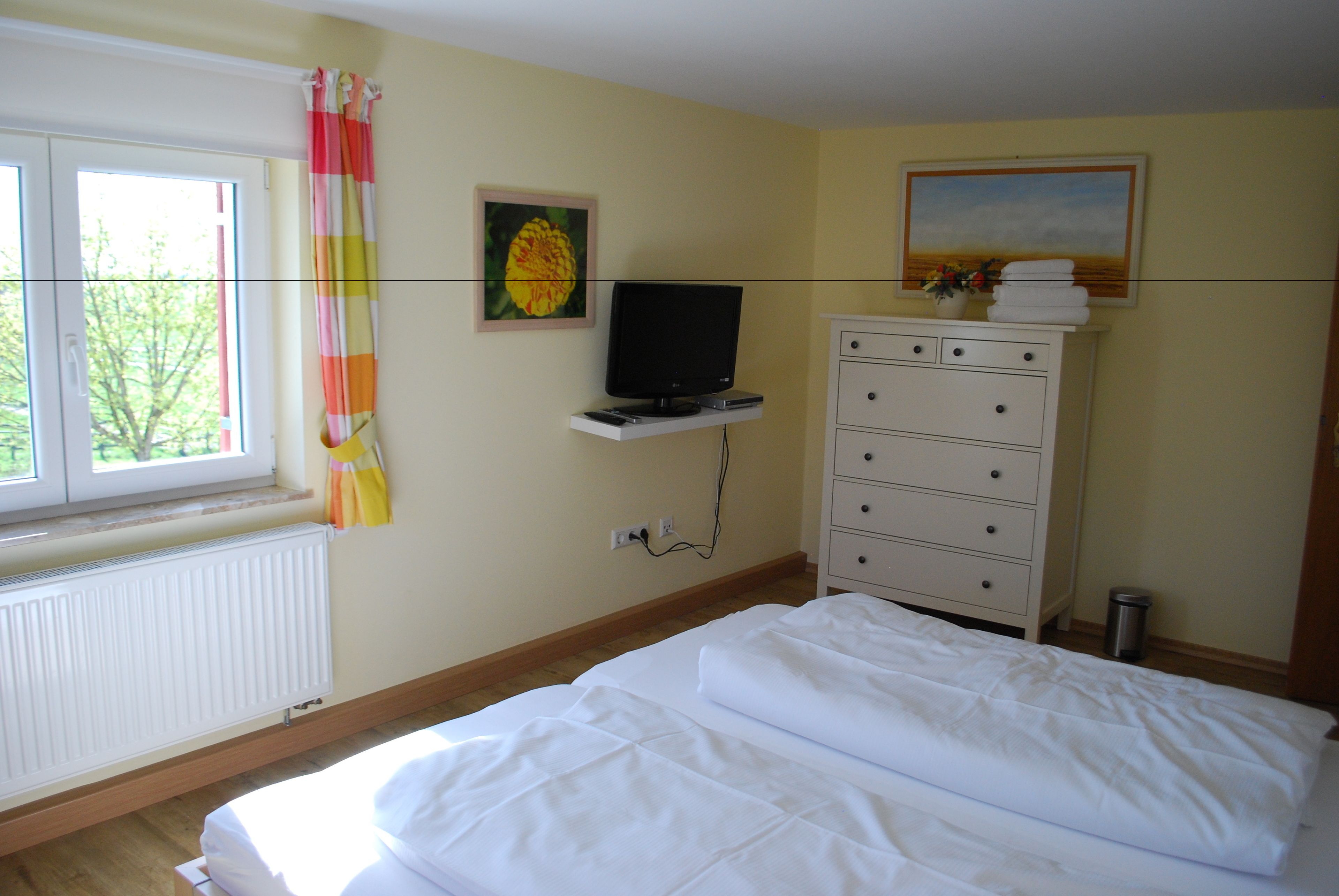 Un dormitorio con cama, cómoda, televisor sobre un estante, radiador y ventana con cortinas.