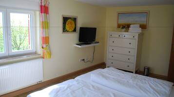 Un dormitorio con cama, cómoda, televisor sobre un estante, radiador y ventana con cortinas.
