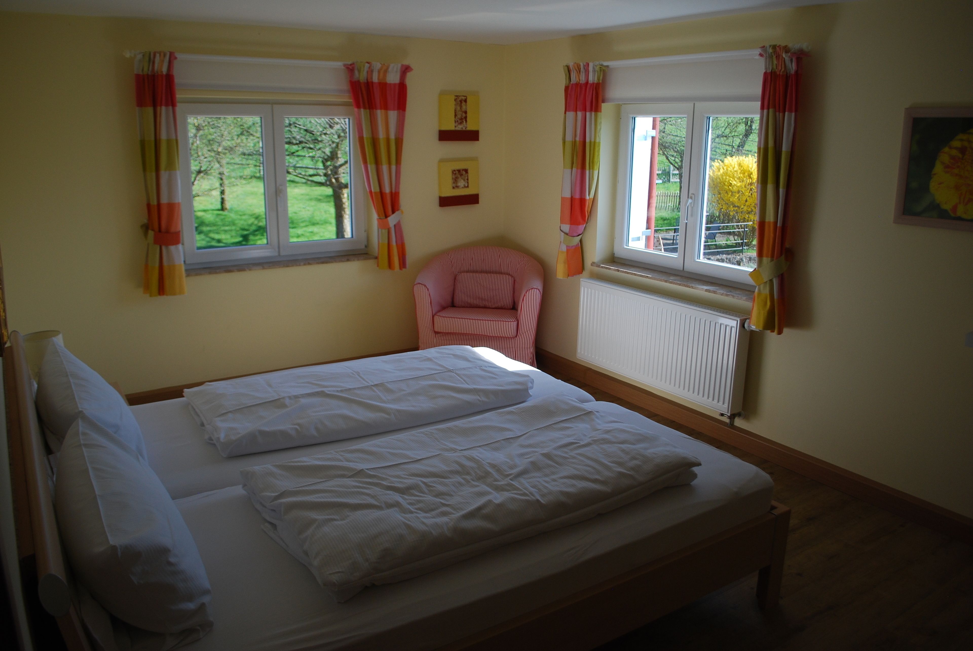 A bedroom with a bed, two windows with curtains, a radiator, and a chair.