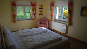 A bedroom with a bed, two windows with curtains, a radiator, and a chair.