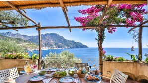 An outdoor dining area with a table set for a meal, overlooking a scenic coastal view with mountains and the sea. - Star Fish House (Ravello)