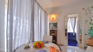 A dining area with a round table, a fruit bowl, a candle, and a magazine. There is a chair, a wall-mounted light, and a view of a blue-tiled room through an open door. - Star Fish House (Ravello)