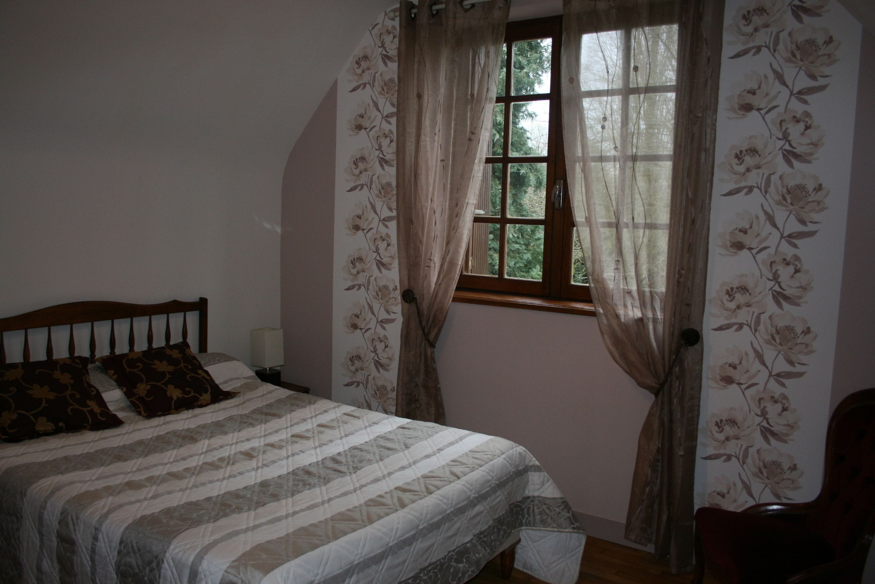 Een slaapkamer met een bed, nachtkastje, stoel en raam met gordijnen.